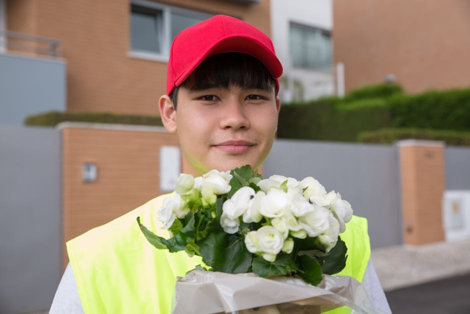 A young delivery person wearing a bright yellow high-visibility vest and a red cap holds a bouquet of white flowers, likely begonias or similar blooms, wrapped in white paper. The flowers are fresh, w