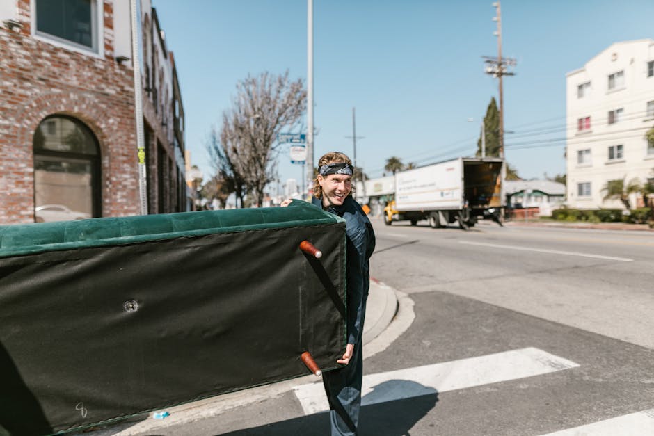 A person with a cheerful expression, dressed in casual dark clothing and a patterned headband, is holding a large, black insulated flower delivery box with red side handles on a sidewalk in an urban a