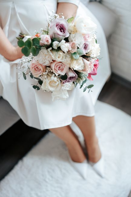 A bride seated indoors on a plush white rug, holding a large floral bouquet composed of pale pink and cream roses, white peonies, and sprigs of baby's breath, accented with eucalyptus leaves. The bouq