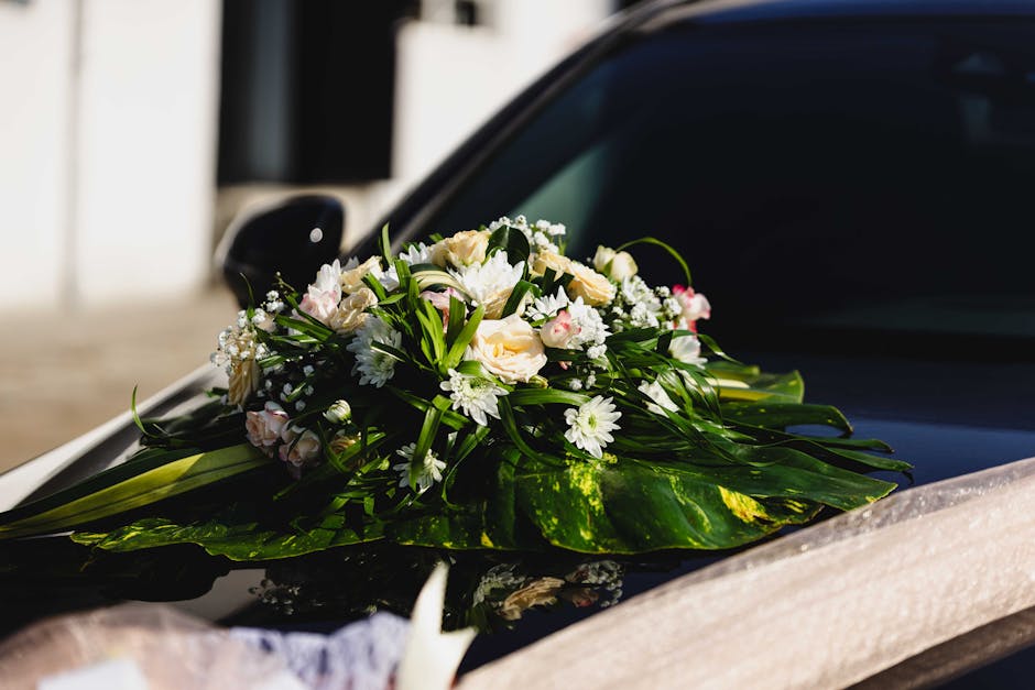 A fresh floral arrangement featuring white roses, daisy chrysanthemums, and delicate baby's breath, accented with lush green foliage, rests on the hood of a black car. The bouquet is carefully wrapped